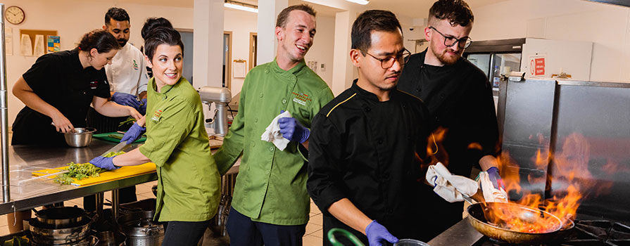 Several chefs are shown working in a busy kitchen. Chefs Mark Paterson and Krista LeTerte are shown wearing green, chef’s jackets in the center of the group. 