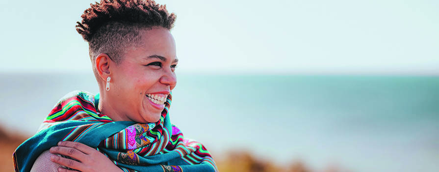 A woman wearing a colourful scarf, smiles broadly while looking towards the horizon. Water can be seen in the background.