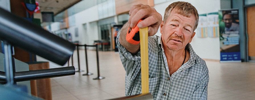 A man sitting in a wheelchair and wearing a buttoned-up plaid shirt and jeans, measures a railing with a yellow measuring tape. 