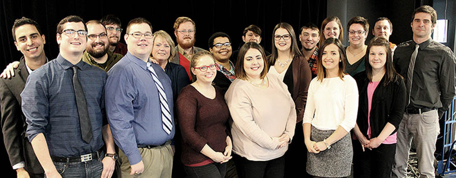 A group of 19 individuals stands close together and smiles for a photo.