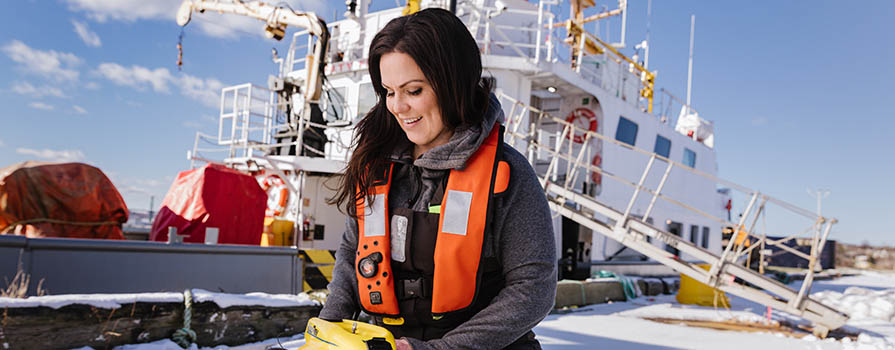 A woman with brown hair is crouched down. She is wearing an inflatable live-vest and holding a small underwater drone that is yellow. In the backgroun, a large, white ship and a gangway can be seen.