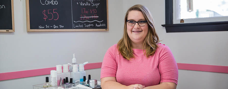 "A young woman in a pink shirt sits behind a desk and smiles. On the desk, nail polish and other aesthetic equipment is seen. There is a chalkboard in the background that reads mani/pedi combo. "