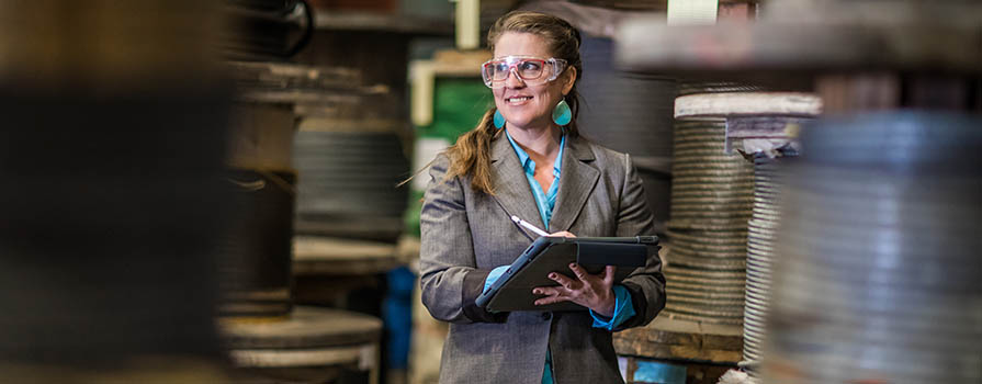 A woman wearing a blazer, safety glasses and large, turquoise earrings can be seen through a frame made by industrial ropes and supplies. She is looking off to the side of the image and writing on a clipboard.