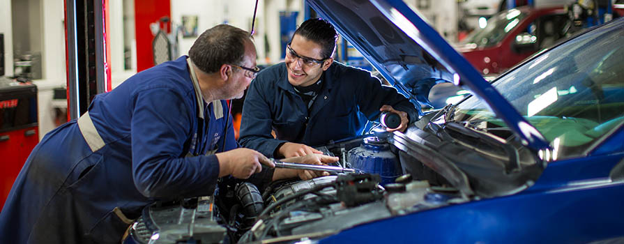 Two men talk while leaning over an open car engine bay. The car and both men's workwear is also blue. They are in an automotive shop.