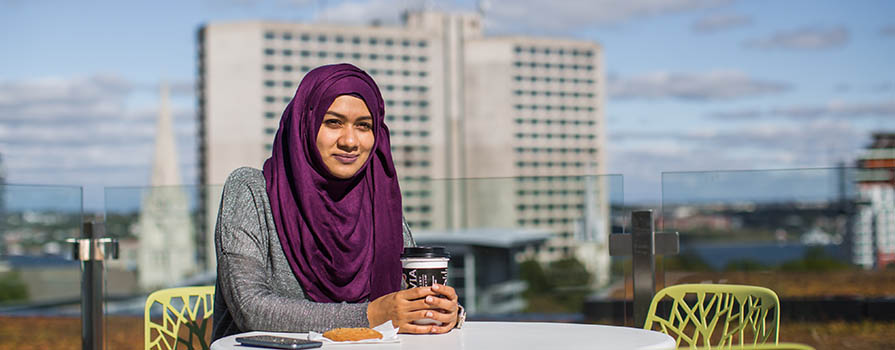 A woman, wearing a purple hijab sits outdoors in a modern green chair at a small round table. In the background tall buildings can be seen. 