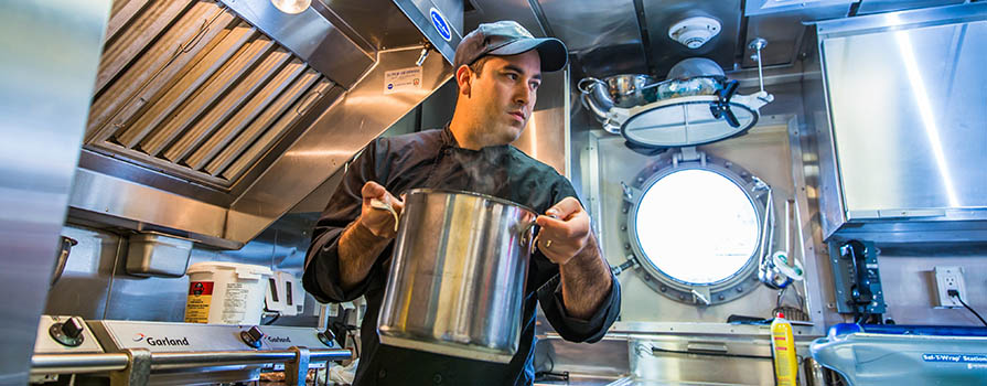 A man wearing a black chef's jacket and black ball cap, transfers a large, stainless steel pot across a kitchen. The kitchen is entirely stainless steel and small. A porthole window is open in the background.