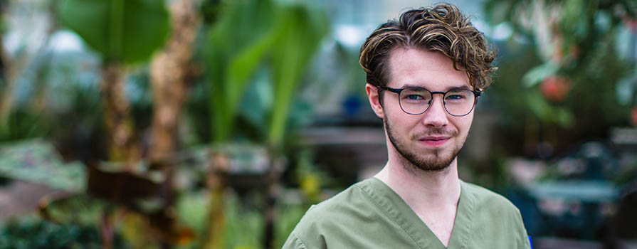 A young man wearing green scrubs and glasses looks into the camera. In the background lush, green plants can be seen.