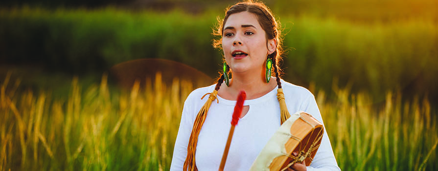 A woman wears a white shirt and holds a pepkwejete’maqn (Mi'kmaq drum). She is singing and beating the drum with a stick with a red end. Her hair is braided. There is a green field behind her.