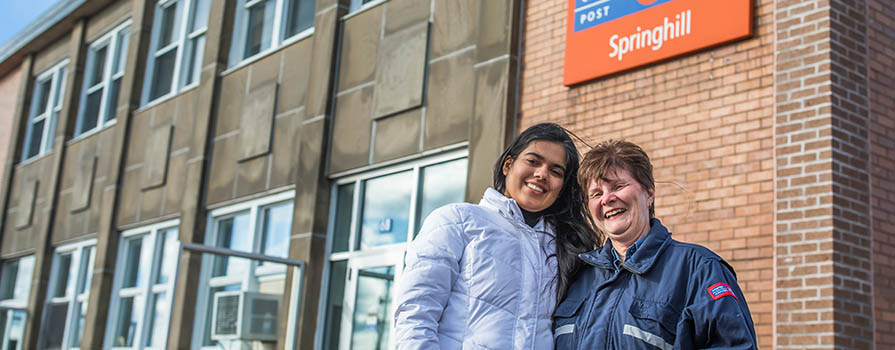 Two women stand in front of a Canada Post Office. One is wearing a white, winter jacket. The other woman is older and wears a Canada Post jacket. 