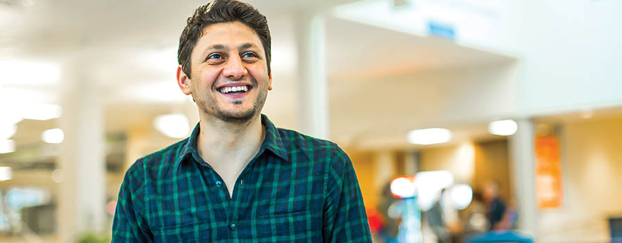 A young man looks off to the top right off the image. He is smiling while sitting at a table. His hand is on a book. He is wearing a button-up plaid shirt. The background is bright and open.