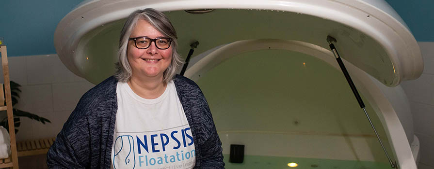 A woman sits on the edge of a large, white flotation pod that is filled with water. She is wearing a sweater over a t-shirt with nepsis written on it. She is smiling. 
