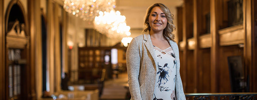 A young woman smiles and looks at the camera. She is wearing a blazer and flowery shirt. She is in a large room with multiple chandeliers, wood paneling and soft seating.