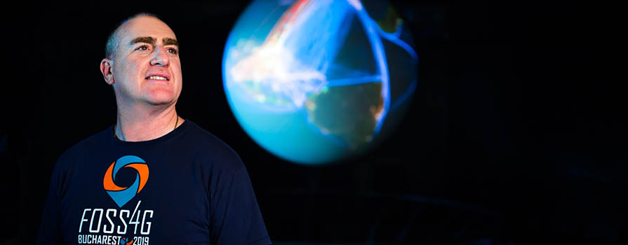 A bald man looks off to the top, right of the image. He is wearing a t-shirt that reads F O S S 4 G Bucharest 2019. Behind him, a well-lit, large globe can be seen.