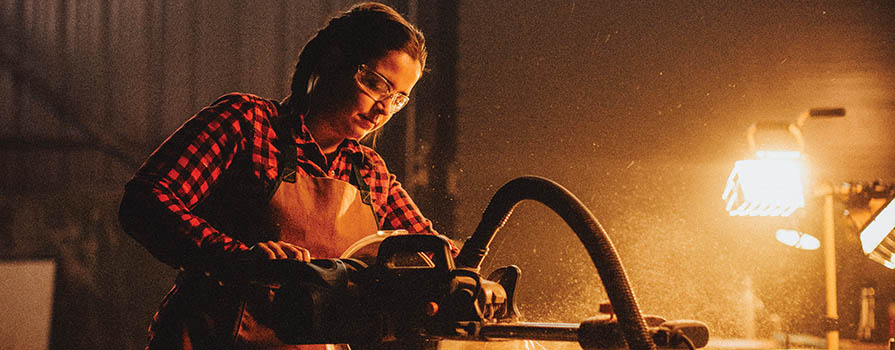 A woman wearing a buffalo plaid shirt, leather apron and safety glasses, cuts wood with a power saw. Sawdust can be seen in the air. There is a light shining in the background.