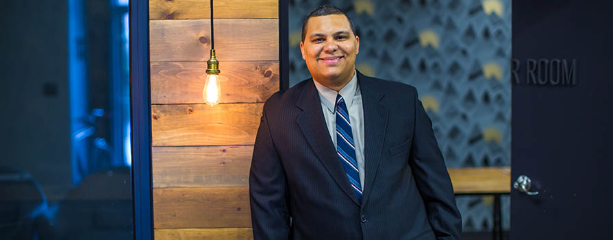 A young man leans against a door casing while smiling at the camera. He is wearing a blue suit and tie. There is a wood clad wall and exposed lightbulb to the left. Behind the man is a room with modern wallpaper.