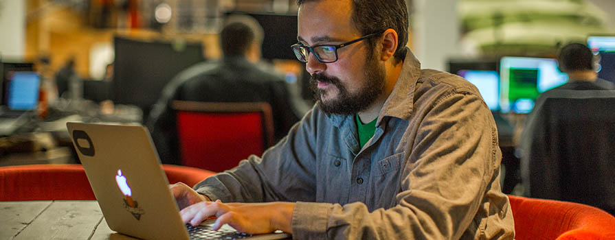 A bearded man wearing glasses sits in a red chair at a round desk and works on an Apple laptop. Behind him, other people doing similar work can be seen.