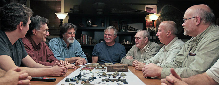 Alex Lagina, Marty Lagina, Rick Lagina, Dave Blankenship, the late Dan Blankenship, Gary Drayton and Doug Crowell all sit around the head of a long table on the set of the History Channel show The Curse of Oak Island. On the table, dozens of pieces of wood, potter, bone and metal are laid out. Most men are looking to Rick Lagina, who appears to be talking. 