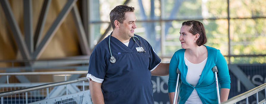 A man in blue scrubs with a stethoscope around his neck, walks down a railed walkway with a woman who is using crutches. 