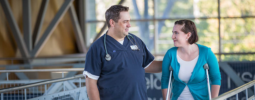 A man in blue scrubs with a stethoscope around his neck, walks down a railed walkway with a woman who is using crutches.