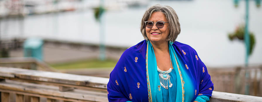 An woman wears a bright, multi-blue-coloured jacket and looks off the left of the image. She is on a wooden walkway and leans up against the railing. Water and boats can be seen in the background.