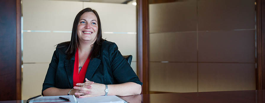 A woman smiles and looks off to the right of the image. She is sitting at a large, brown table and wears a dark blazer and red, v-neck shirt. There is an open notebook on the table in front of her.