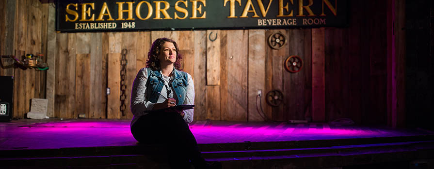 A woman sits on a stage wearing a jean vest and white shirt. She is looking off to the right. The stage wall is clad in rough lumber and a sign reads the seahorse tavern. A pink-purple light is shining on the floor. 
