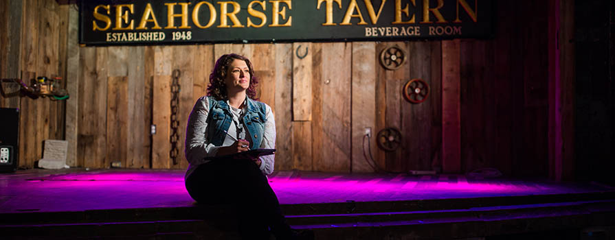 A woman sits on a stage wearing a jean vest and white shirt. She is looking off to the right. The stage wall is clad in rough lumber and a sign reads the seahorse tavern. A pink-purple light is shining on the floor.