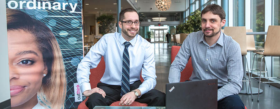 Two young men sit on chairs and look at the camera. They are wearing buttoned up shirts. On a low, glass table in front of them is a black laptop. There is a pull-up banner to the left that reads: IBM outthink ordinary.