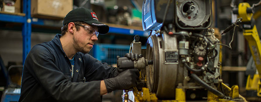A man in blue coveralls, a ball cap, safety glasses and black gloves works on a mechanical item. He is in an industrial shop environment. 
