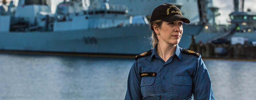 A woman, wearing a Canadian Navy uniform and black cap, looks off to the right. In the background is water and two navy ships.