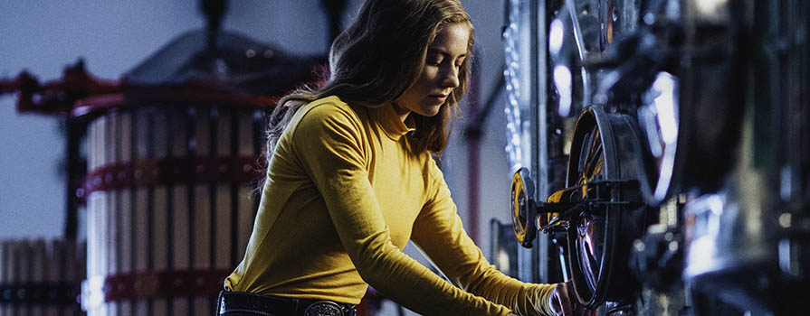 A young woman in a yellow shirt crouches in front of commercial wine-making equipment, which she is operating. The room is dark, and the woman’s reflection can be seen in the floor. In the background, a large, wooden barrel is seen.