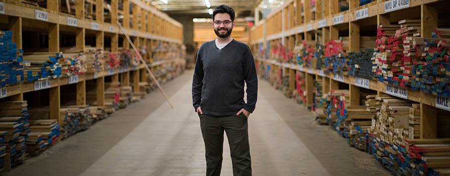 A young man with a beard and glasses, wearing a v-kneck sweater and white t-shirt, stands in the center of an isle in a warehouse. The shelves hold many kinds of wooden planks.