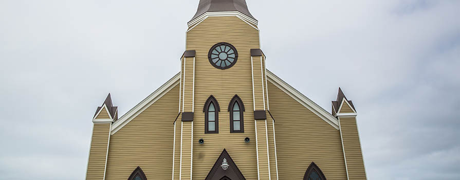 The top half of St. Mary's Polish Parish is seen against a cloudy sky. On the center steeple a metal cross can be seen. A bird is flying on the left.