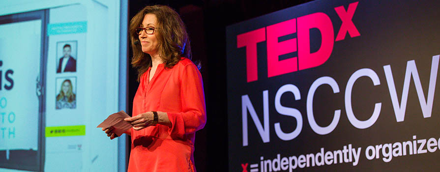 A woman in an orange shirt and glasses stands in front of a large sign that reads TEDx NSCC Wa…(the rest is unseen). She is holding cue cards in her hands and appears to be giving a presentation.