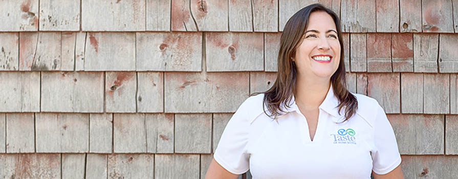 A woman in a white t-shirt smiles while leaning against a wooden shingled wall and looking to the right. On her shirt, the word taste can be read. 