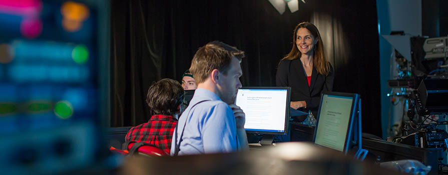 Three individuals are shown in a television studio. It is dark. Two are seated at computers with their backs to the camera. A third person, standing, is looking at the two seated people and smiling. 
