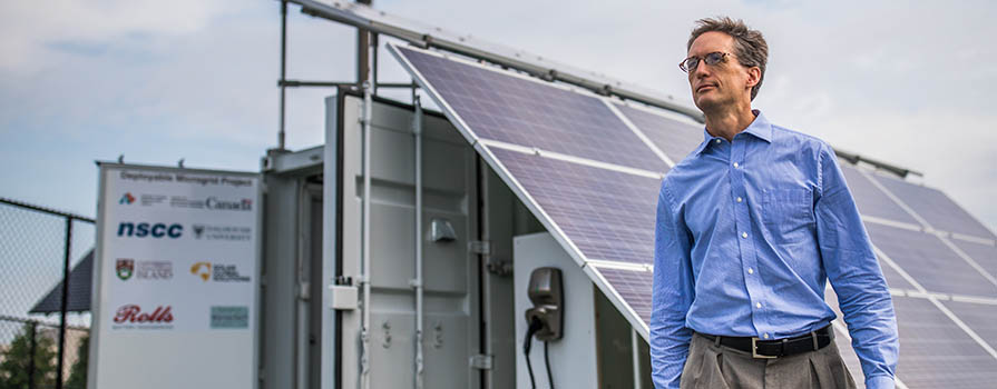 A man in a blue, buttoned up shirt and khaki pants walks in front of a large section of solar panels that are attached to a white shipping container. He is outside.