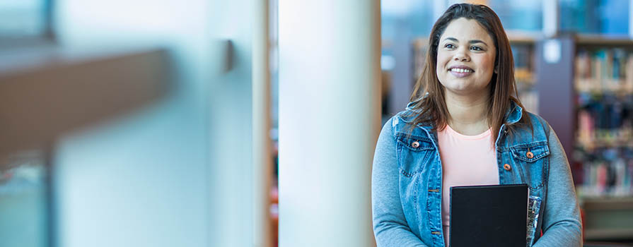 A woman in a jean jacket, light pink shirt and flowery bottoms looks out a window to the left of the image. She is holding a black book to her stomach and smiling. Bookshelves can be seen in the background.