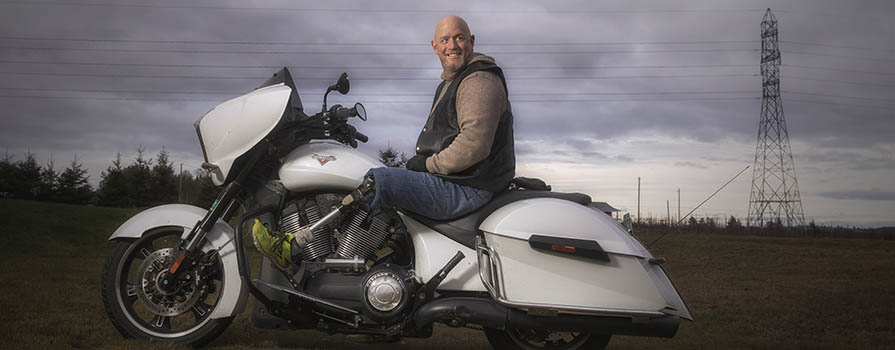 A man smiles while sitting astride a white motorcycle. He is wearing jeans, a brown sweater and a vest. His leg is a prosthetic, and he is wearing a bright green sneaker. There is a large power line in the background.