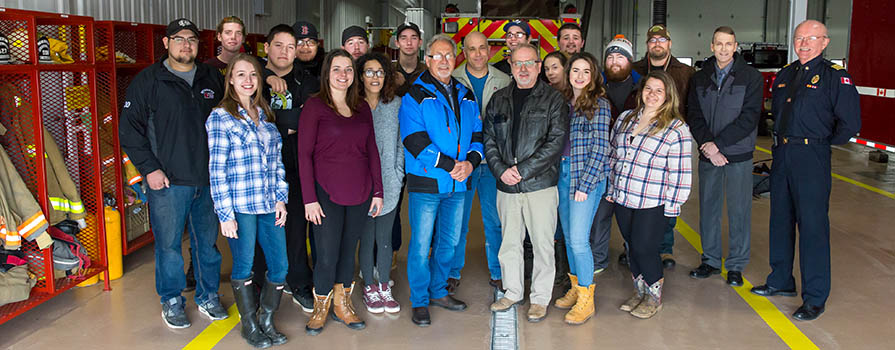 A group of 21 individuals stands close together and smiles for a photo in a firehouse. While most appear to be students, the two on the far right are not. Both are dressed in business attire. One is dressed in a uniform.