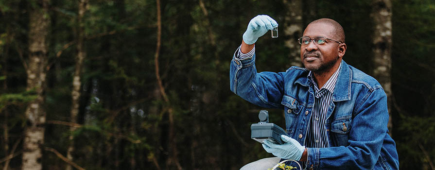 A man crouches by a well. He is wearing a jean jacket and blue latex gloves. He is holding a test tube and looking at it. In the background there is a thick crop of trees.