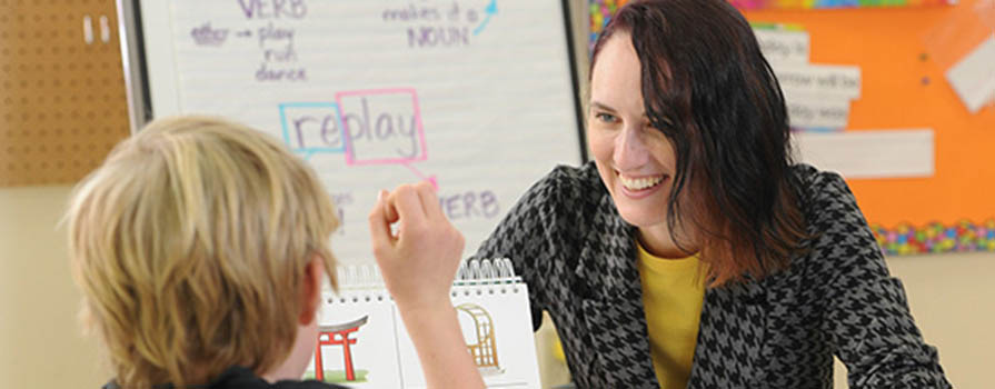 A woman with brown hair sits across a table from a blonde child. The child's face is not shown. The woman is smiling and wearing a yellow shirt and plaid jacket. Behind the woman is a paper pad on an easel and brightly coloured paper attached to a corkboard.