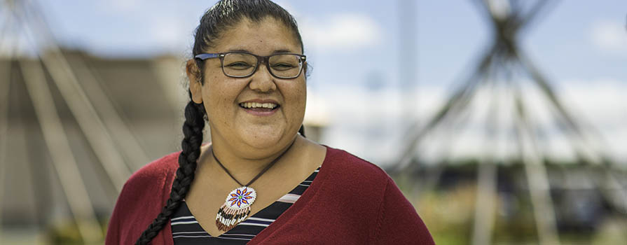 A woman in glasses smiles while looking off to the right of the image. She is wearing a maroon, button up sweater, a black and white striped t-shirt and a beaded necklace. He hair is in a braid. In the background, two teepee frames can be seen. 