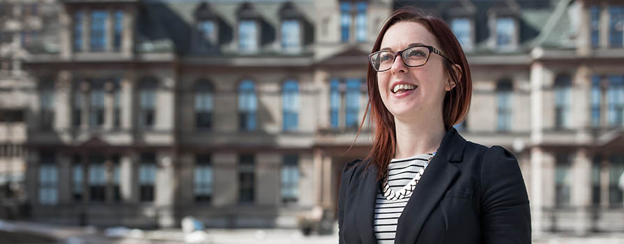 A young woman stands in front of a large, sandstone, townhall building. She is smiling and looking to the top, left of the image. She is wearing a black blazer and a black and white striped t-shirt. Larger buildings can be seen behind the townhall.