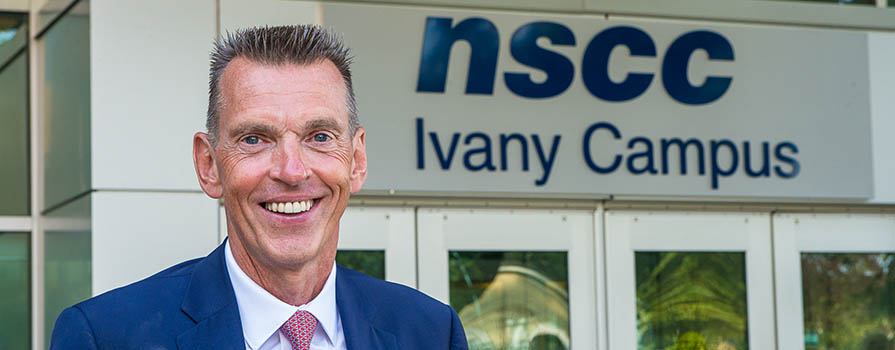 A man, Ray Ivany, stands in front of the doors to NSCC Ivany Campus. He is wearing a blue suit jacket, white shirt and pink tie. He is smiling.