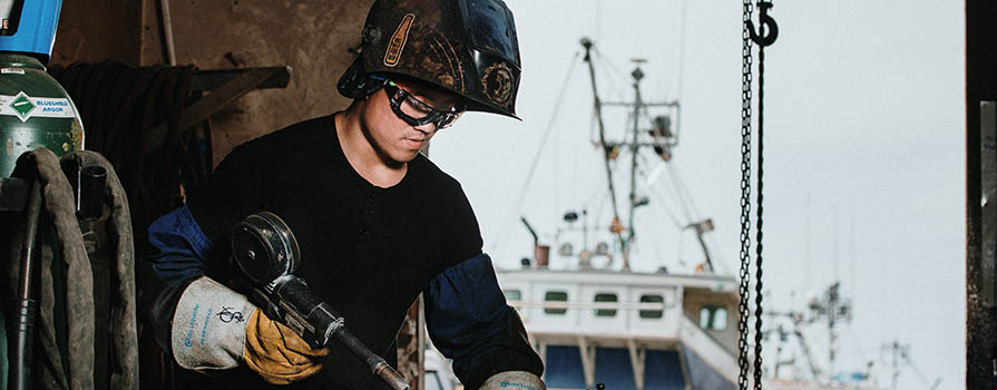 A young man welds a component of a ship. He is wearing a welding helmet (which is open), safety glasses, long leather gloves and a black shirt. In the background, several ships can be seen on land.