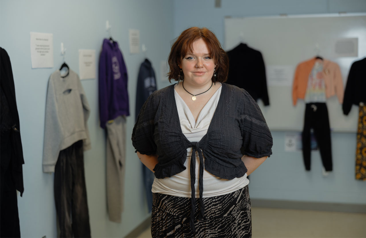 NSCC student Mia stands in the classroom, in front of her exhibit with clothing hung on the walls behind her.