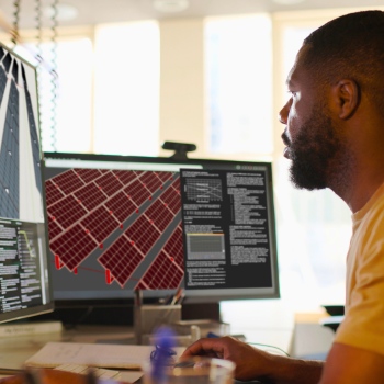 A man engrossed in work at a desk, examining solar panel designs and data on dual computer monitors.