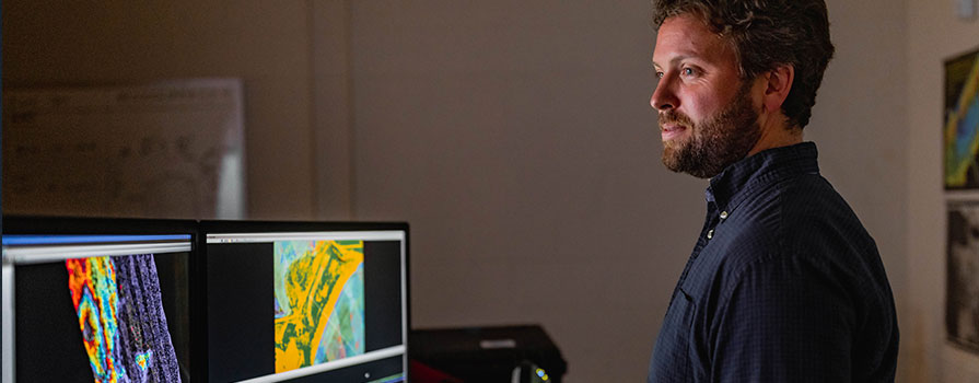 NSCC researcher Nathan Crowell looks at a computer screen in a darkened room. The screen features a colourful, mostly yellow and blue, technical image of Port Hilford Bay. The image was created using data collected on the bay’s seafloor, tidal current velocities and flushing rates.