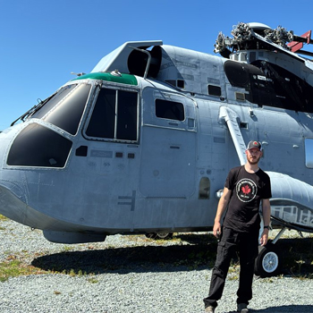 Matthew stands outside, in front of a military helicopter. 